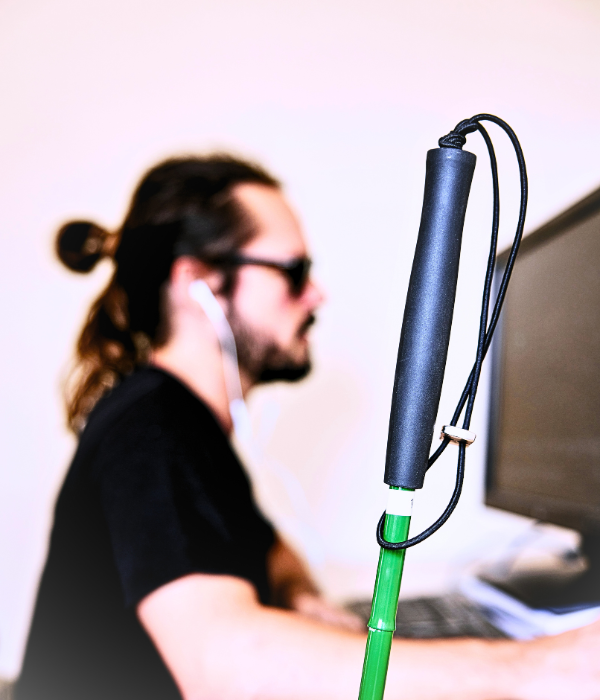 Man in sunglasses working on a computer with a white-tipped cane in the foreground